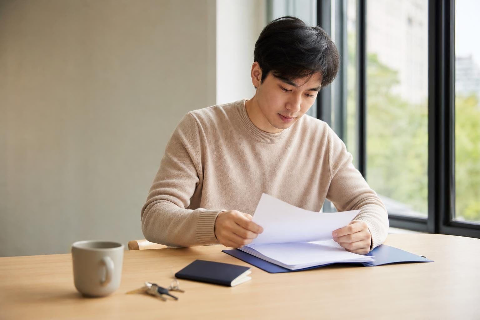 A man at a wooden desk reading blank papers from a folder beside a passport, keys, and a mug